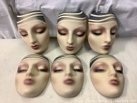 Front view of six full face mannequin masks with painted feminine faces and striped headbands arranged in two rows of three on a white surface.