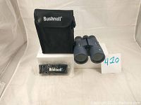 Binoculars next to black Bushnell branded soft case and cleaning cloth, shown on white box with lot number card.