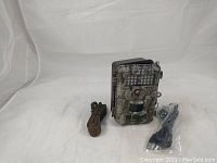 Front view of Bushnell Trophy Camera Camo Model 119447 with night vision LEDs, lens, motion sensor visible, strap and USB cable placed next to it.