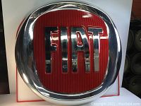 Front view of large circular FIAT emblem with red background and chrome FIAT letters inside a chrome frame.