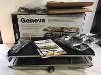 Full view of SwissMar Geneva Raclette Grill box, grill, instruction manual, and additional accessories packed in plastic wrapping on top of the unit.