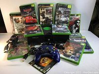 Front view showing the original Xbox console, blue wired controller, power and AV cables, and 13 game cases displayed upright and lying flat