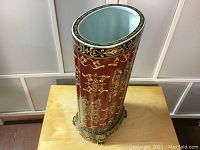 Front full view of the red patterned oval ceramic umbrella stand with brass base and feet on a wooden surface.