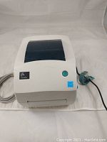 Top view of the Zebra GK888t label printer showing the white unit with a dark blue transparent cover and green power button. Power and USB cables attached.