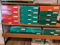 Multiple file storage drawer units stacked in three columns on a wooden shelf; colors include red, green, orange, and white. Each unit has a front white pull handle and label slots with white paper labels.
