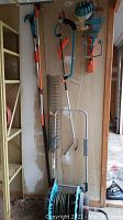 Gardena garden tools including long-handled pruner with orange and silver handle, hedge shears or trimmer and metal rakes hanging on wall above hose reel
