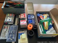 Photo showing boxes with colorful paper clip holders, marker packs, boxes of staples, label sticky notes, protractor sets in cases, compasses, and ink cartridges on black desk protector.