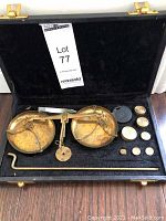 Open black case showing brass precision balance scale with two pans, central beam, and chains, alongside assorted brass weights and a rod on a velvet-like lining.
