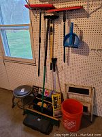 Wide view showing multiple brooms, dustpan, bucket, step stool and long-handled cleaning tools hung on pegboard wall and standing on floor.