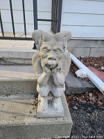 Front view of weathered concrete gargoyle statue sitting on outdoor step, showing facial expression and texture.