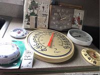 Overview of six vintage clocks and thermometers arranged on a counter, showing large Cooper thermometer among other clocks and thermometers