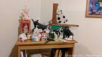Photo of multiple pig and cow figurines on a wooden table including boxed Enesco Country Pig, wooden decorative cow sign, piggy bank with hearts, and assorted ceramic figurines.