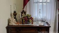 Wide shot showing all items on wooden sideboard including etched glass decanter, etched glasses, amber oil lamp, religious figurines and grapes.