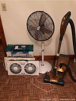 Photo showing three white dual-window fans, Holmes stand fan, and Kenmore vacuum cleaner placed on floor against wall.