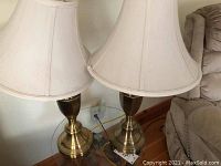 Pair of brass-like table lamps with beige fabric bell-shaped shades shown on wooden floor beside beige couch.