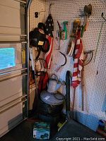 Photo of garage wall pegboard displaying garden tools, string trimmer, blower, flag, watering can, metal garbage can, and other items.