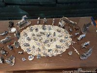 Photo of multiple miniature metal animal figurines arranged on a wooden table with a lace doily in the center. Various animals including horses, deer, birds, lion, fox, and small animals.