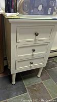 White wood nightstand with three drawers and metal knobs, standing on tiled floor.