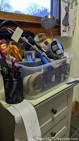 Plastic tote container filled with various office supplies including scissors, pens, packing tape, desk lamp, and laptop bag on top of a cabinet.