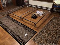 Photo of three runner rugs laid on parquet flooring, one rug shows black border and tan diamond lattice pattern, some household items and metal bed frame parts also visible.