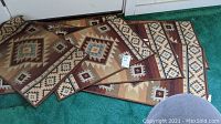 Seven Southwestern style throw rugs arranged piled overlapping on green carpet, showing brown, tan, cream, blue and red geometric patterns.