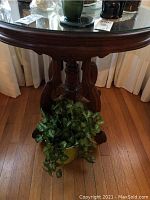 Full view of round wood side table with clear glass top, carved pedestal base, and curved legs. Plant placed underneath the table.