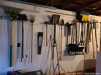 Wide view of yard tools hanging on pegboard including rakes, saws, shovels, pitchfork, spade, and snow shovels