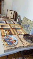 Table display showing six boxed collectible plates with certificates, three framed sets of prints, a welcome glass hanging sign, and a small dried flower framed piece