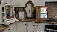 Four mirrors displayed on a kitchen counter: a large wooden framed mirror, ornate gold framed mirror, ornate uniquely shaped gold frame mirror, and a distressed white framed mirror with shelf