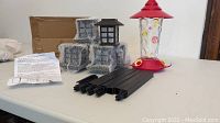 Hummingbird feeder with glass container, red top and base with yellow flower feeding ports, and six solar light stakes and lamps wrapped in bubble wrap on table with instructions.