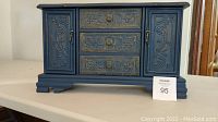 Front view of the large blue painted jewelry box with ornate carved drawer fronts and two side doors closed.