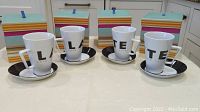 Four white latte cups with black letters L, A, T, E on the front, each paired with a black and white saucer, displayed in front of their striped decorative boxes