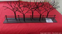 Metal tree sculpture showing multiple stylized bare trees with branching limbs along a fence row on a flat base against a red background.