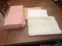 Seven folded tablecloths on wooden table, showing variety in color and texture: pink, gold, white, and white lace.