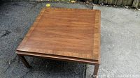 Photo of the solid wood square coffee table showing the top surface and legs from one angle on asphalt floor outdoors.
