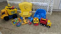 Photo showing an assortment of colorful toddler toys including a smiling yellow Fisher Price chair, blue booster seat, toy trucks, and puzzles on the floor with white safety gate in background.