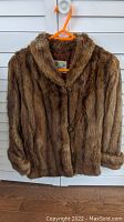 Front view of the brown mink fur jacket hanging on an orange hanger against a white shuttered door, showing button details and collar damage.