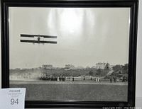 Full framed black and white photo showing the bi-plane flying over a large field filled with spectators and buildings in background.