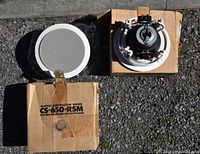 Pair of Klipsch CS-650-RSM ceiling-mounted speakers with one showing the front grille and the other showing the back with wiring and mounting hardware on cardboard boxes.