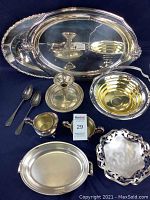 Full view of assorted silverplated servingware pieces arranged on dark cloth, including trays, bowls, spoons, creamer, and candle holder.