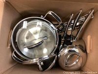 Box containing various stainless steel pots, pans, and lids with some surface wear visible on items.