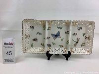 Front view of rectangular porcelain plate showing butterfly, dragonfly, bee, and flower painted designs with pierced gold-accented border and black stand.