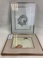 Two framed prints shown together: conch shell print in silver frame leaning against white background, lady kneading bread print in wood frame laying flat.