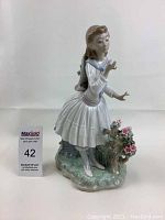 Front left side view of the Lladro porcelain figurine depicting a girl in white dress with blue scarf and pink flowers on base.