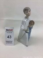 Front view of Lladro figurine showing boy holding candle and child clinging to him, with MaxSold lot number card adjacent.