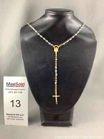 Blue bead and gold-tone metal rosary necklace displayed on black neck form, showing full length and cross pendant.