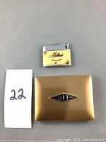 Front view of cigarette box and vintage lighter placed on a neutral grey surface with lot number card