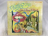 Front cover of Steely Dan Can't Buy A Thrill record album, showing colorful surreal graphic design with lips, teeth, a banana, and people on a street.
