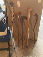 Five wooden walking canes and sticks leaning against a grey wall next to a cooler in a garage.