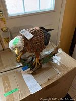 Photo of eagle sculpture showing white head, yellow beak and talons with brown body perched on branch with green leaves, on a cardboard box.